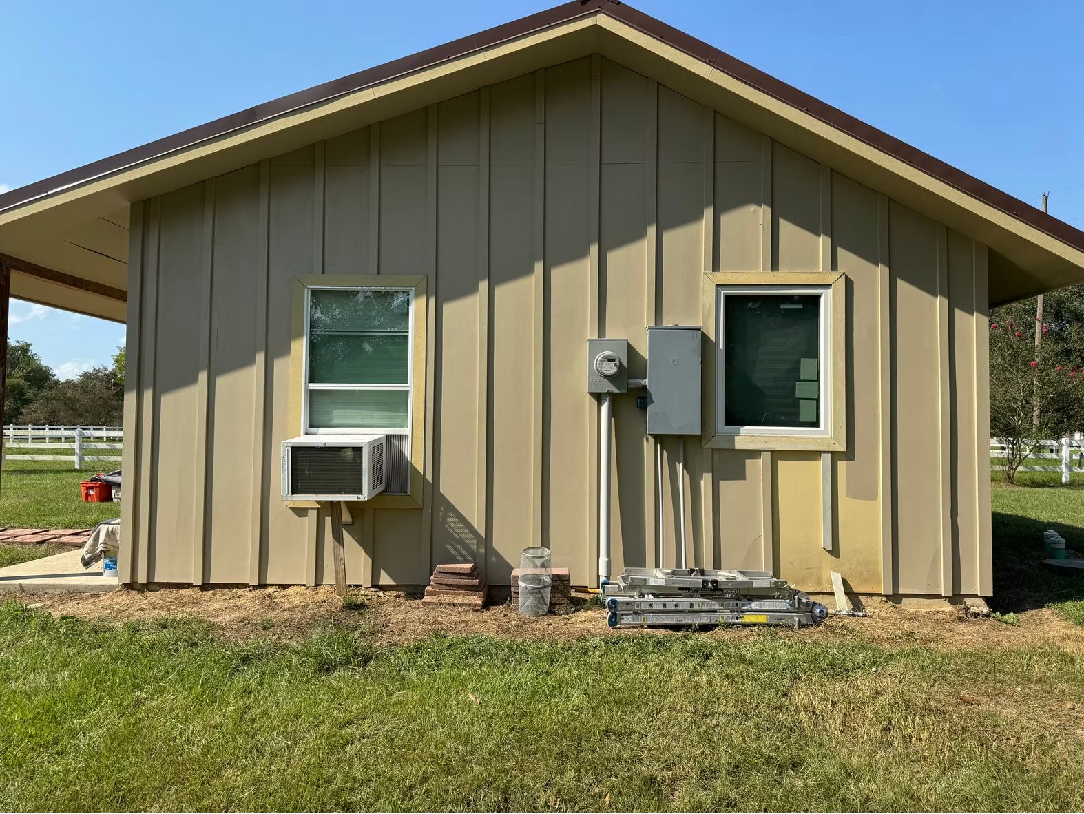Small modern residential building with tan metal siding and two windows, featuring a window air unit and electrical panel