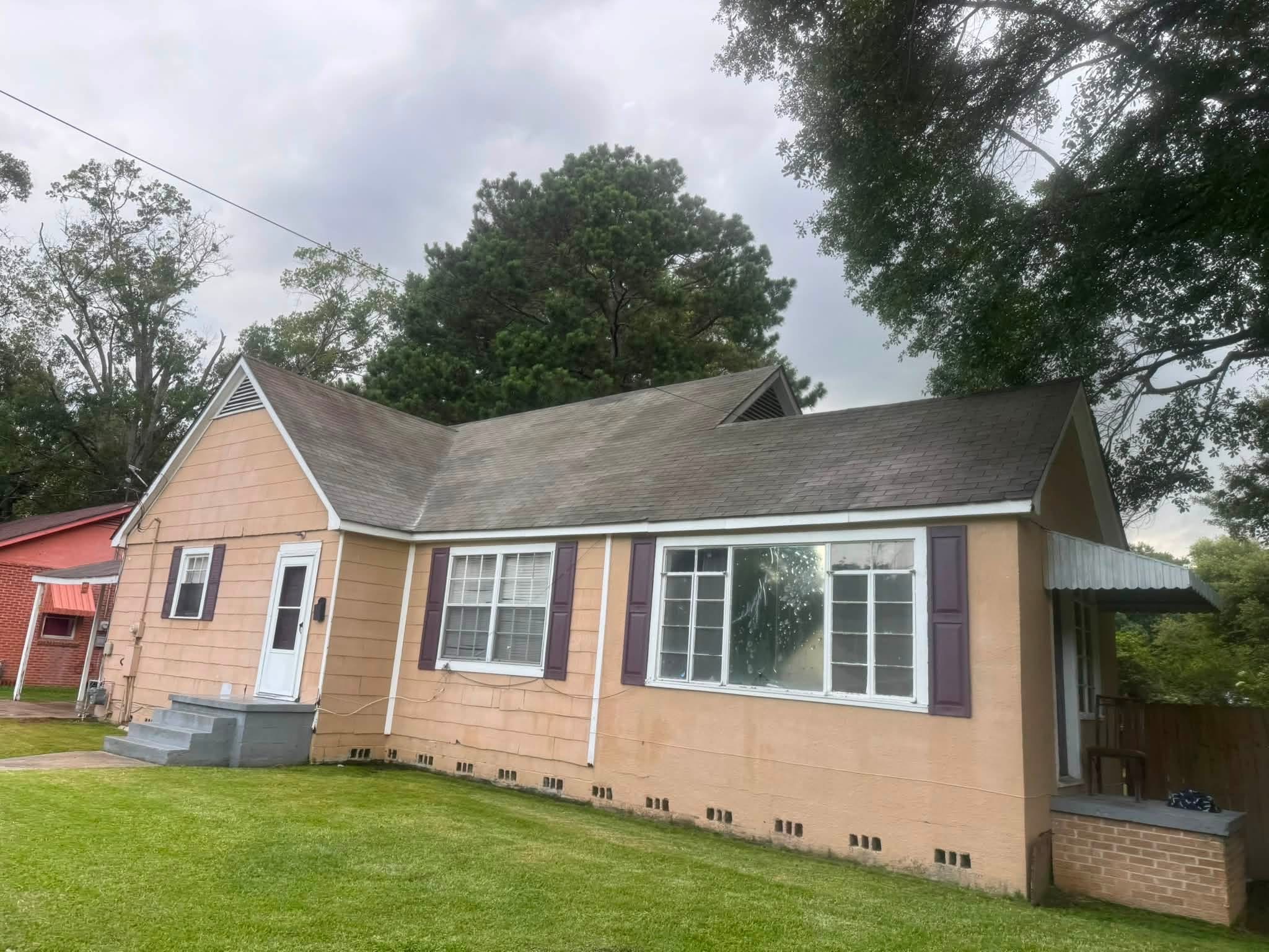 Single-story tan house with purple shutters and gray roof surrounded by trees and green lawn